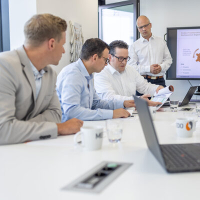 Geschäftsleute diskutieren Projektunterlagen in einem Meetingraum, im Hintergrund eine Präsentation auf dem Bildschirm.